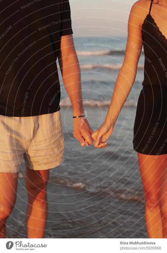 Unrecognizable couple holding hands on the beach. Man and woman together with waves of sea on background. Closeup framing. Tanned skin at sunset. Love and relationships. Summer love story of young adults. Millennial on vacation.