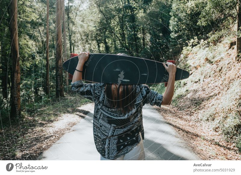 Young woman from behind walking with long board in the middle of the forest. boho carefree enjoyment excitement females freedom hippie horizontal laughing