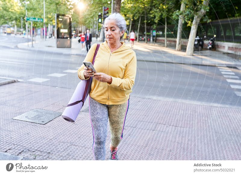 Aged sportswoman walking on street and using smartphone training fitness social media sidewalk online female senior aged elderly mat sportswear vitality