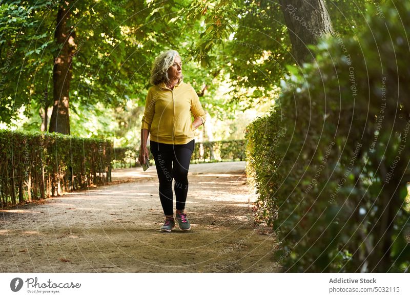 Aged sportswoman walking in park path training cardio fitness summer activity alley female senior aged elderly bush shrub lush athlete stamina physical workout