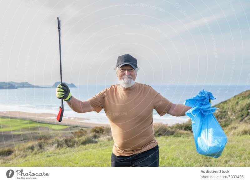 Positive senior man standing on hill with trash tongs and sack pensioner smile volunteer garbage environment nature collect sea pollute rubbish ecology waste