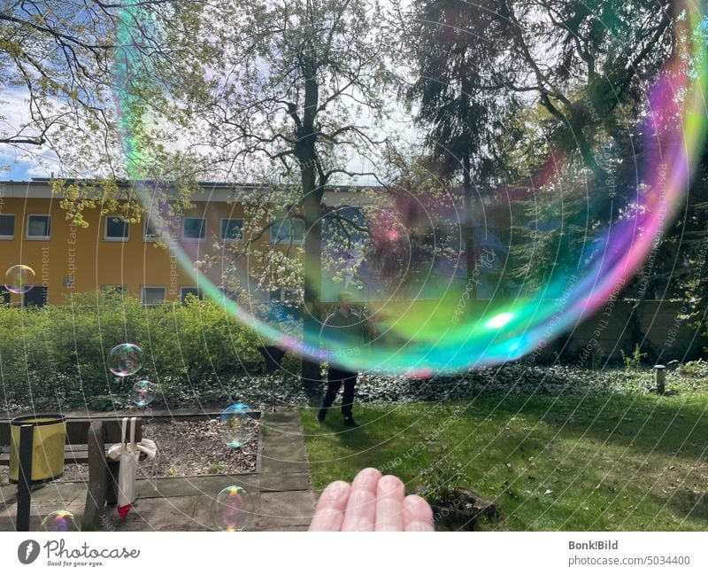 A person lets fly soap bubbles. In the foreground a hand and a huge silk bubble, shimmering in rainbow colors, slowly floating away. In the background, others that have already flown a distance. Garden, playground.