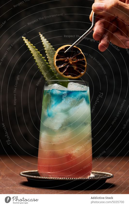 Anonymous person preparing rainbow paradise cocktail garnished with leaves and orange fruit alcohol drink glass Rainbow Paradise leaf beverage aperitif booze