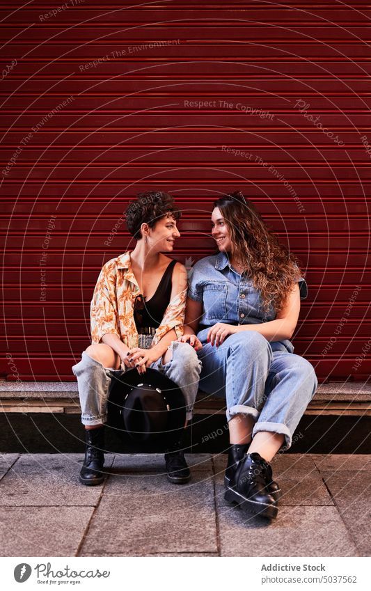 Lesbian couple sitting near red wall women smile date girlfriend border happy building street madrid spain female young together friendship optimist positive