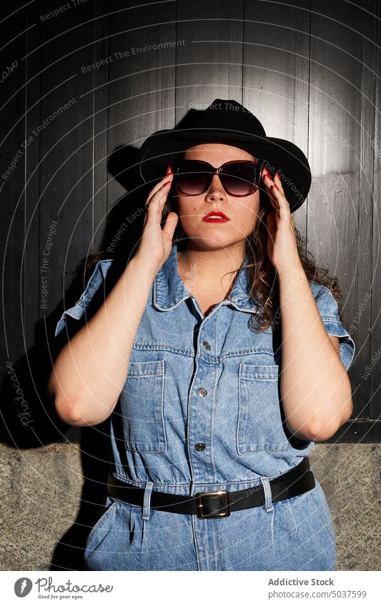 Stylish young woman touching sunglasses style adjust appearance wall hat casual confident portrait madrid spain female eyewear personality denim street