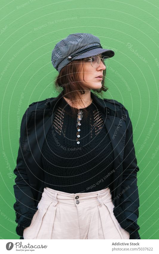 Stylish woman against green wall style outerwear street urban appearance modern individuality building female young outfit glasses cap hand in pocket accessory