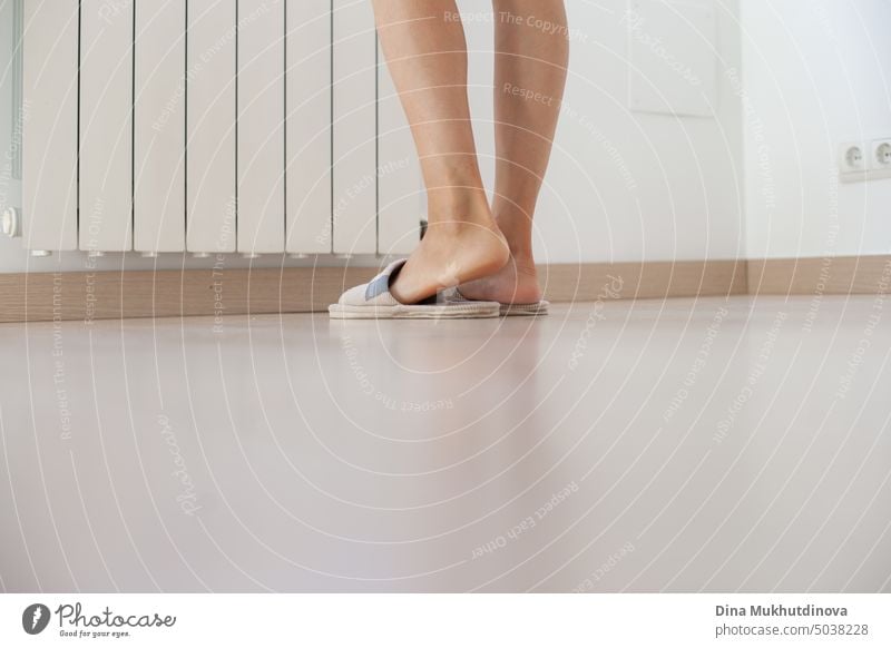 Woman standing near heater in the apartment at home, wearing slippers. Cold winter in Europe. Legs and feet closeup. Neutral beige color palette. Gas prices for heating homes. Energy crisis.