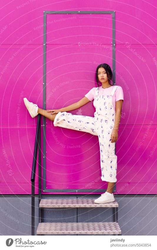 Asian woman looking at camera with serious expression, raising one leg over railing near pink wall. asian chinese fashionable urban japanese person girl female