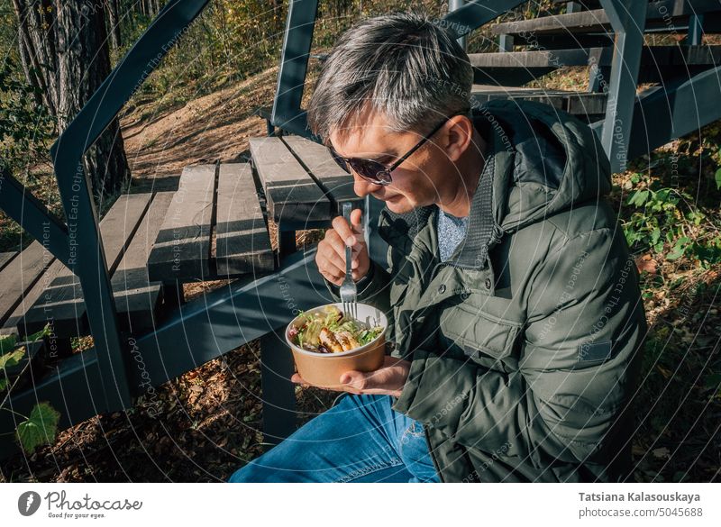 A man dines on a salad from a disposable paper bowl in the open air during a walk in nature in the fall park lifestyle food lunch take away eat outdoors adult