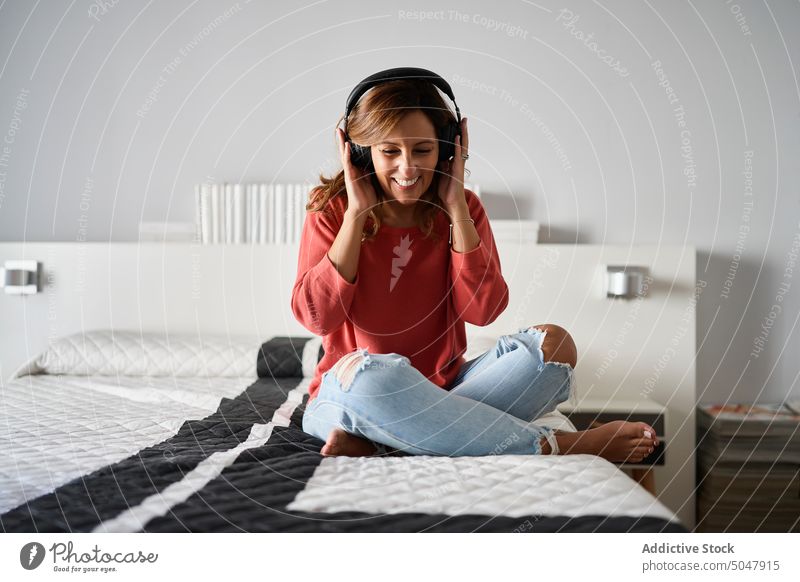 Woman listening to music on bed woman home weekend bedroom headphones touch meloman female hispanic ethnic middle age mature casual legs crossed looking down