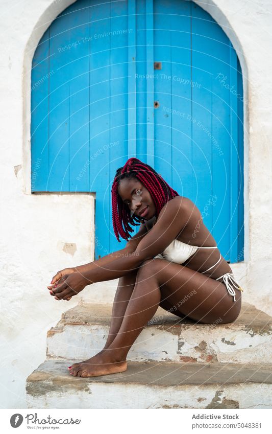 Stylish woman sitting outside house with blue door step tourist bikini summer vacation relax sensual dreadlocks outfit confident trendy young exterior entrance