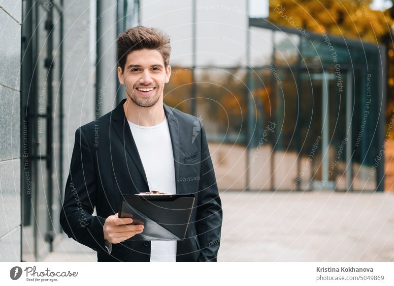 Portrait of young handsome man in jacket with documents, utility bills, report. Businessman near office building. He is satisfied with work of corporate staff.