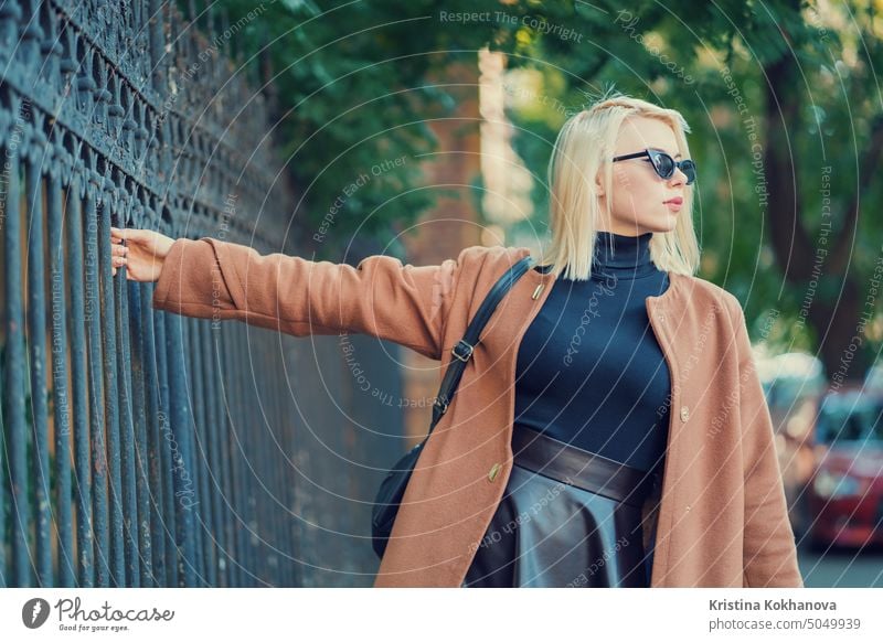 Mysterious stylish blonde girl walks through the European metropolis, holding the old fence. Portrait of young woman in fall fancy look. Traveling concept city