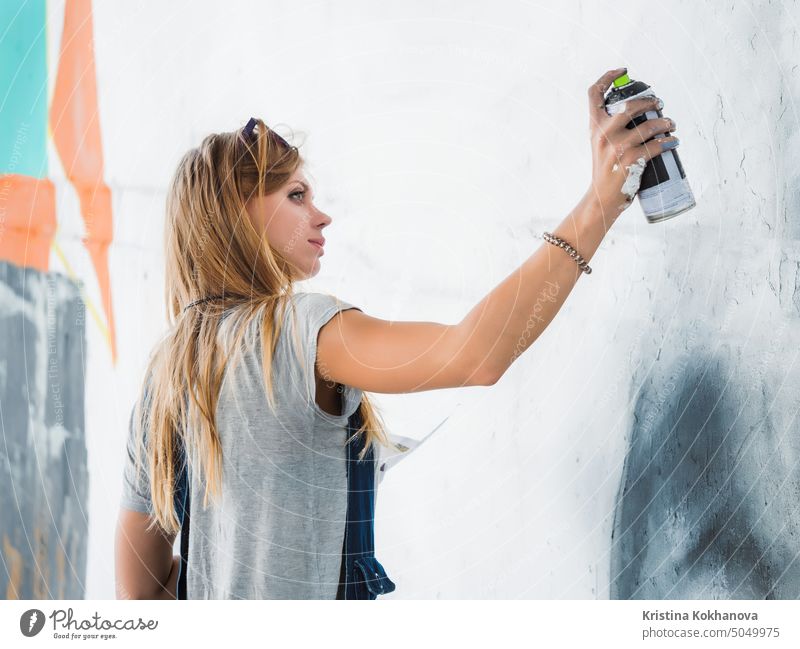 Beautiful Young Blonde Girl making graffiti of big eye with aerosol spray on urban street wall. Creative art. Talented student in denim overalls drawing picture
