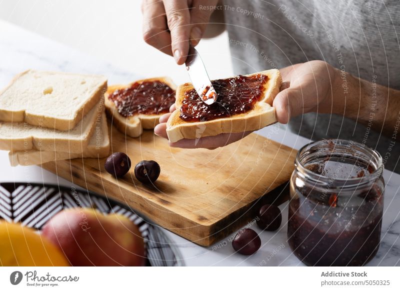 Crop man preparing toasts with cherry jam prepare breakfast spread knife smear food delicious sweet bread meal male morning tasty cook table fresh yummy kitchen