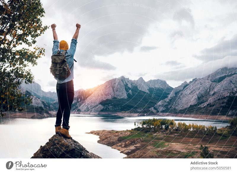 Traveler standing on rocky formation near lake backpacker traveler tourist fists up raised hands mountain peak achieve observe admire trip journey man male