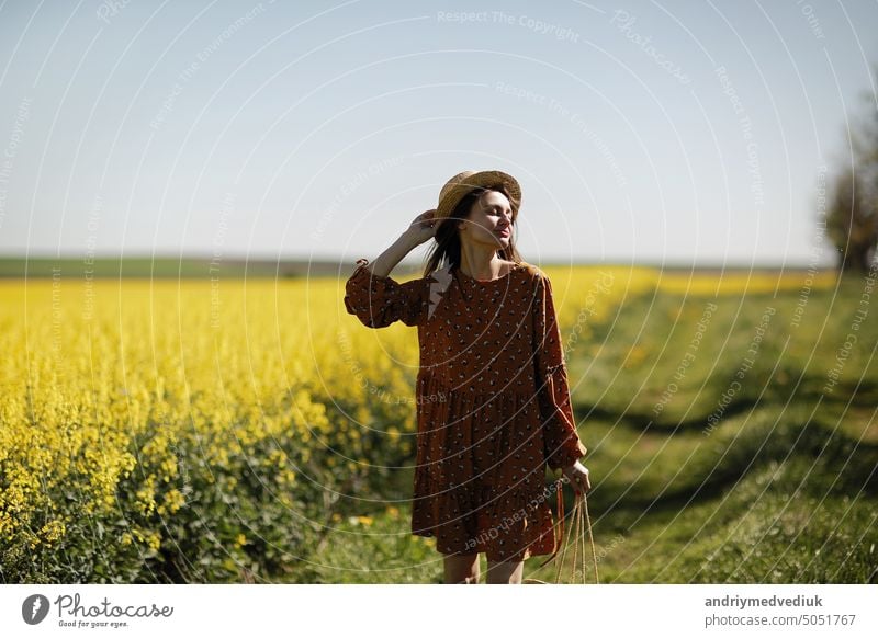 beautiful young woman walks in a field of yellow rapeseed. Girl brunette long hair fly in wind dressed in dress and straw hat. summer holiday concept female