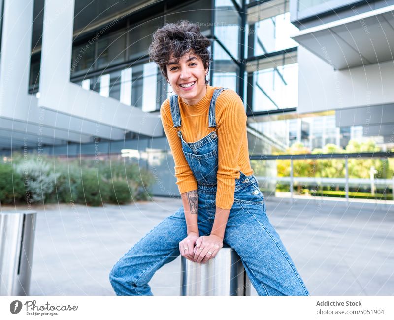 Positive lady sitting near building woman street cheerful summer city enjoy smile style chill jeans female urban positive brunette overall casual denim hispanic