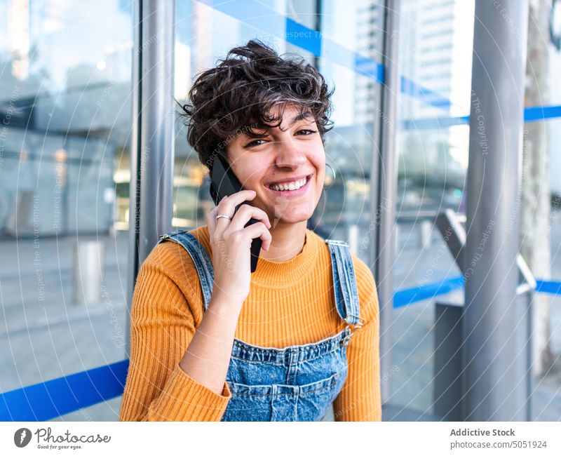 Cheerful woman talking on phone near modern glass building cheerful smile smartphone phone call street urban glass wall mobile female speak conversation happy