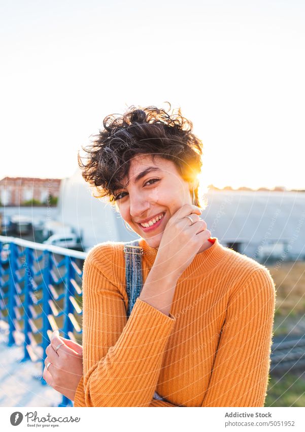 Cheerful woman with short hair standing on footbridge smile positive fence touch face happy sun barn cheerful young female sweater casual daytime optimist