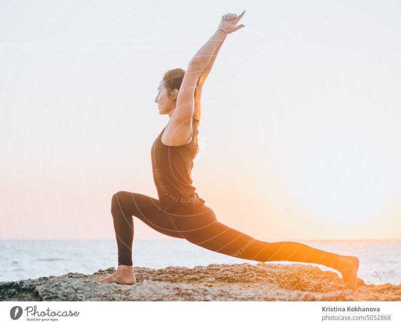 Slim woman in black bodysuit practicing yoga near sea or ocean during sunrise light. Flexibility, stretching, fitness, healthy lifestyle. abstract active adult