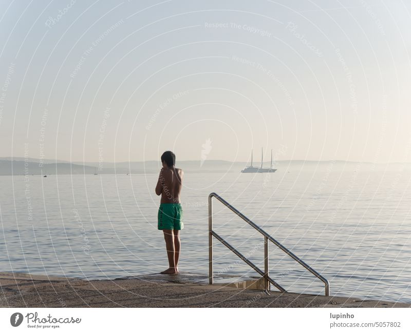 A boy standing on the city beach of Trieste Boy (child) November Afternoon Ocean Adriatic Sea vacation Light hazy ship Green tranquillity Italy Smooth