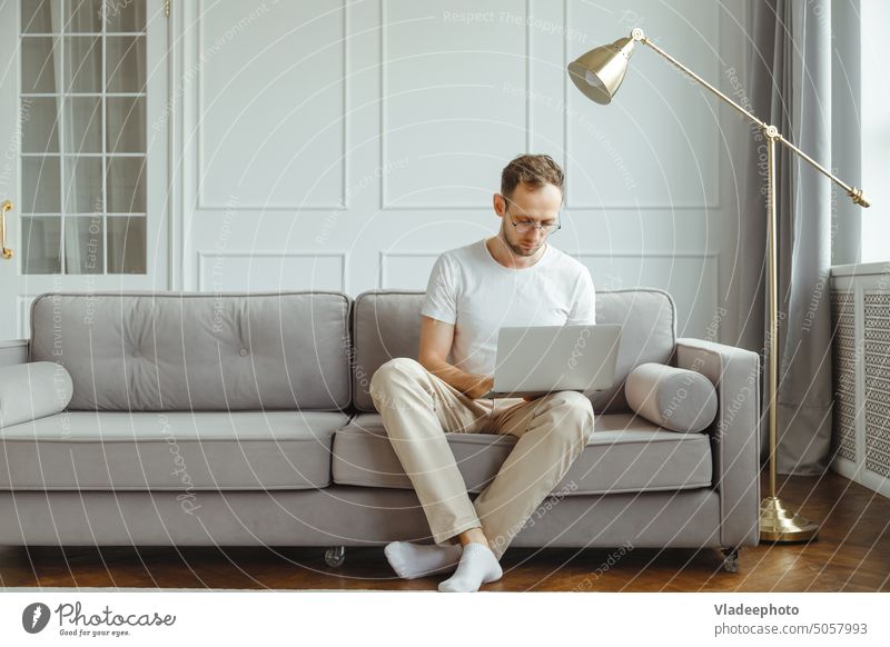 Young European man freelancer sits on comfortable couch in living room, works on laptop Sofa Man Home freelancers Technology Computer Easygoing Couch masculine