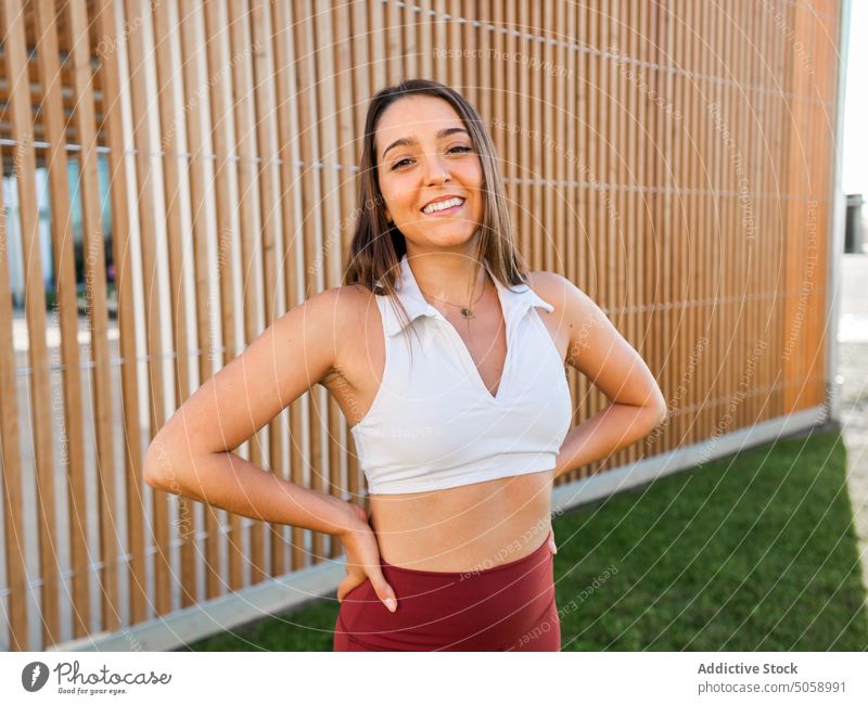 Cheerful sportswoman standing near wooden fence smile hand on waist sportswear athlete positive park grass healthy break female young workout confident wellness