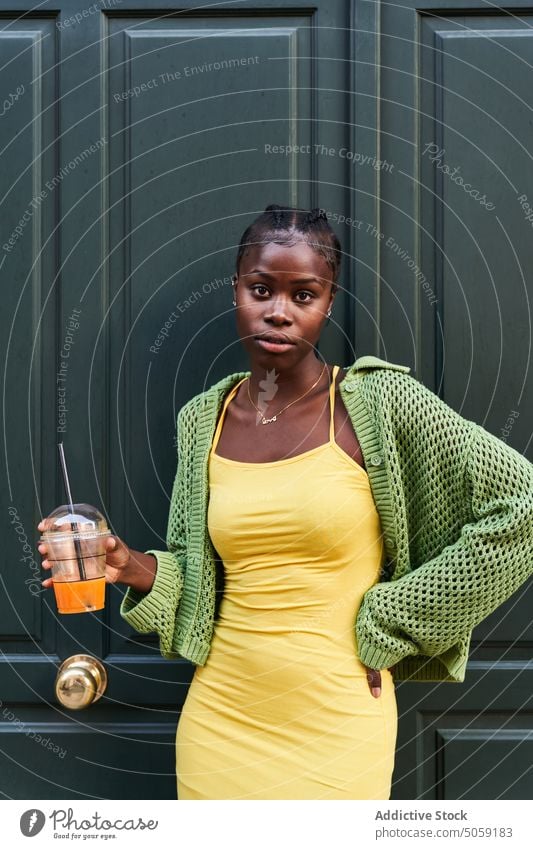 Stylish young black woman with cup of takeaway juice standing near door style confident street hand on waist serious portrait city drink refreshment female