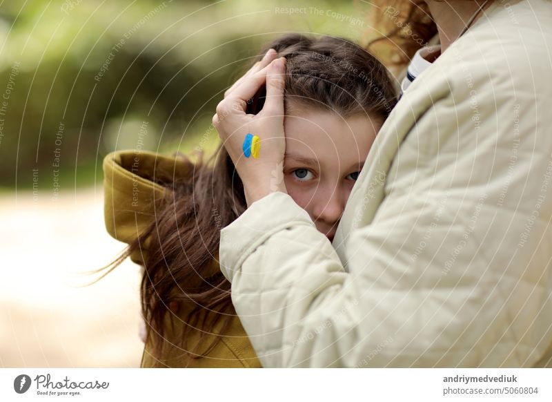Close up of caring young mother cuddling sad little daughter with flag on hand outdoors. Peace, no war, help, stop russian aggression. Invasion of Russia in Ukraine. High quality photo
