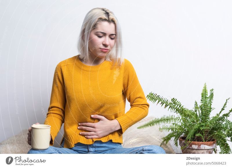 Blonde woman sitting on ground holding cup of tea and one hand on belly blonde nausea coffee person indoor pain abdomen illness female problem stomach adult