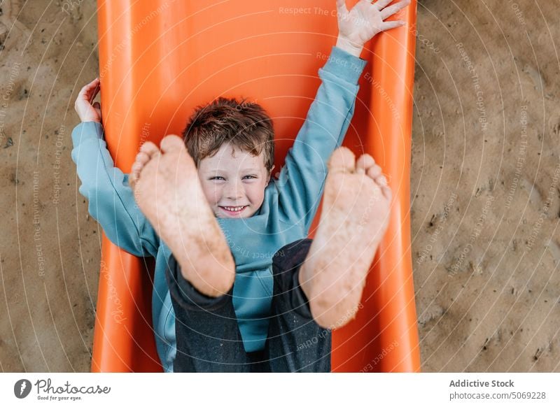 Merry boy playing on orange slide playground sand barefoot show dirty happy weekend summer preteen kid pastime arms raised smile glad childhood positive