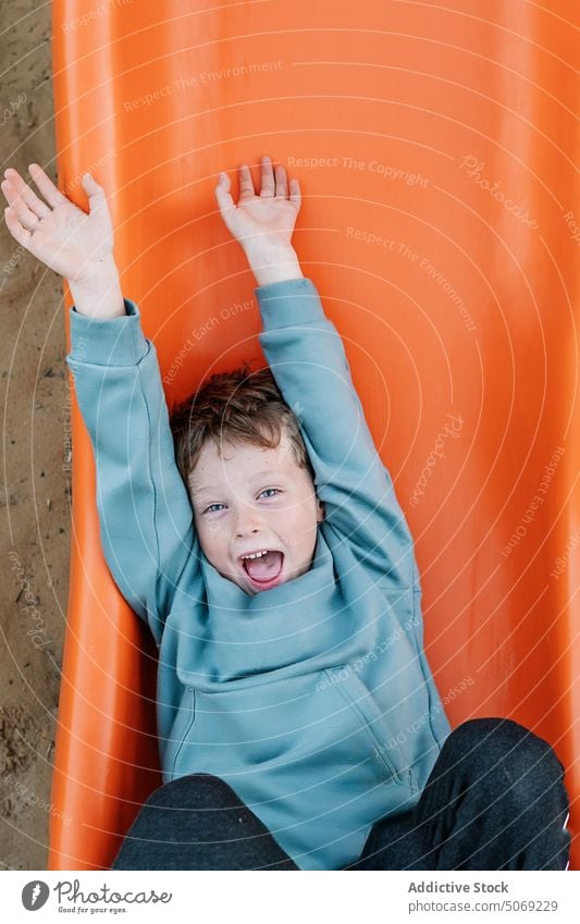 Merry boy playing on orange slide playground sand barefoot show dirty happy weekend summer preteen kid pastime arms raised smile glad arms stretched childhood