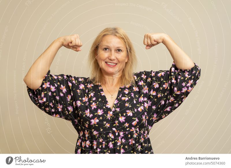 Caucasian blonde woman in a dark patterned dress raising her arms and flexing her biceps muscle isolated over beige background. Strong and brave modern woman concept.