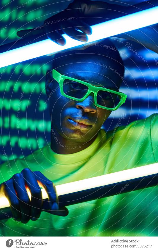 Cool trendy black man in sunglasses touching neon lamps in disco club cool metaverse emotionless extraordinary fashion goggles luminous fluorescent male glow