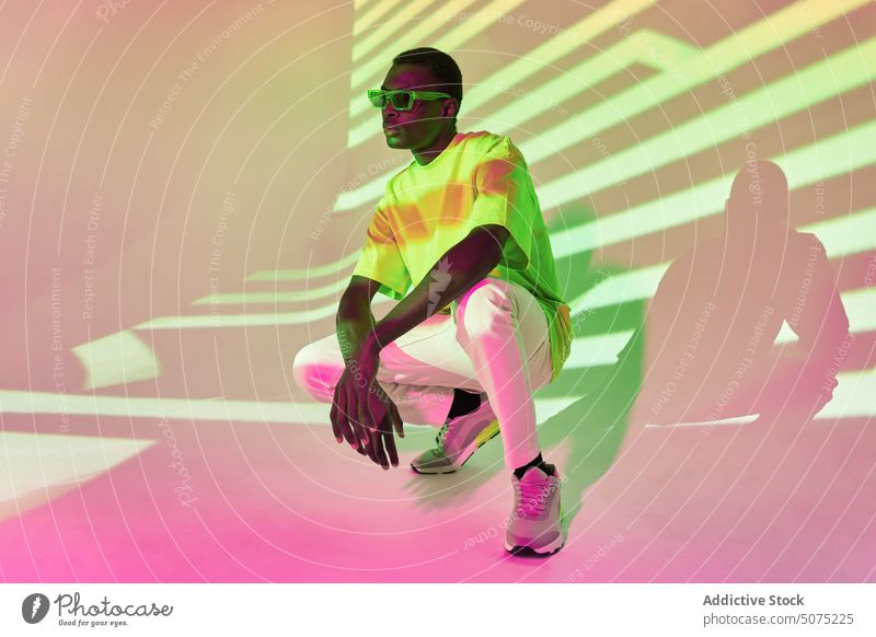 Cool black man in green T shirt and sunglasses posing in shadows crouch down cool african effect pose stripe casual metaverse confident style bright light male
