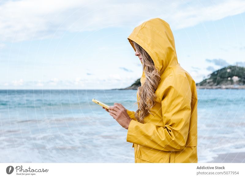 Woman browsing smartphone on beach woman seashore coast ocean raincoat surfing internet mobile female text message yellow spain connection sms using free time