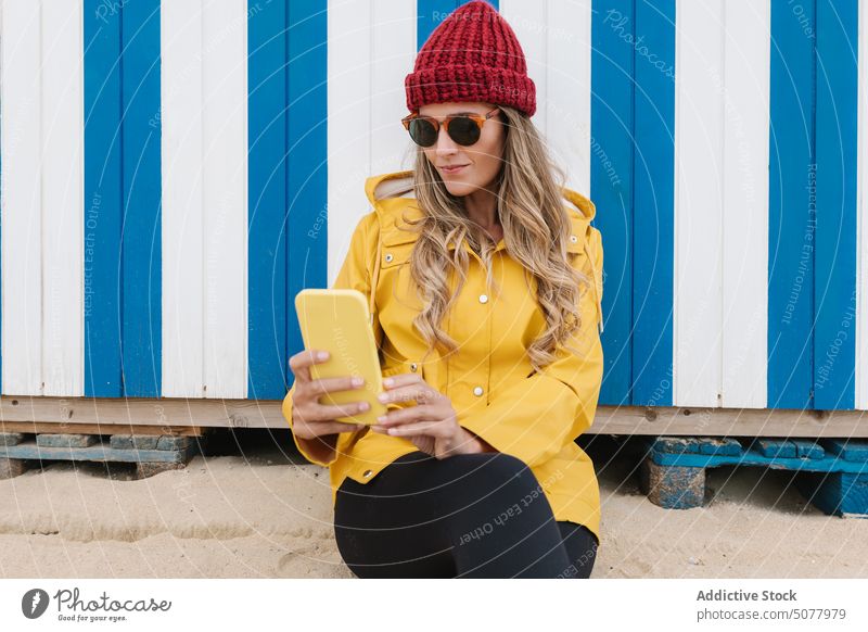 Fashionable woman in sunglasses and raincoat taking selfie on beach smartphone style seashore colorful memory travel scroll trendy blond mobile moment donostia