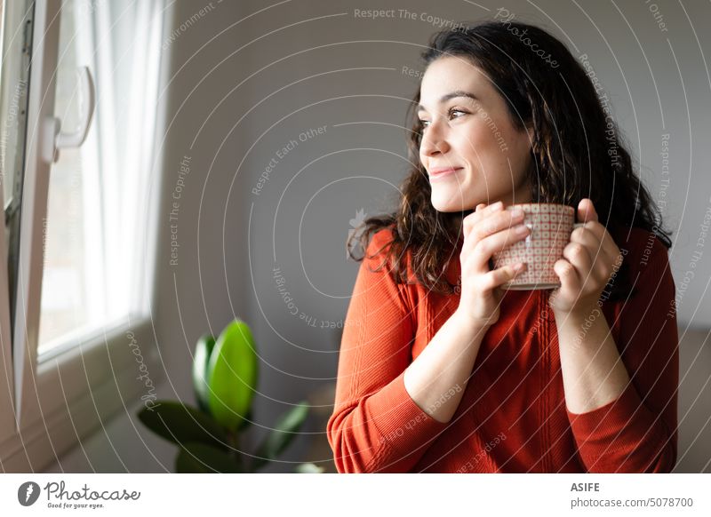 Young happy beautiful woman holding a cup of hot drink and looking trough the window home coffee tea smiling looking away relaxed sweater orange red comfort