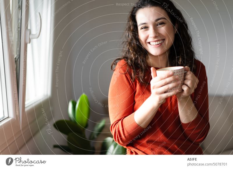 Happy young beautiful woman having a coffee or tea at home happy smiling laughing looking at camera toothy holding cup drink window relaxed sweater orange red