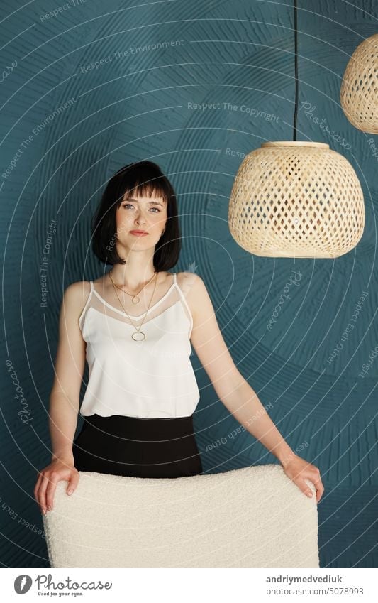 Millennial young brunette woman looking at camera and standing in creative office with grange blue green wall, soft chair and rattan lamps . Successful self-confident female designer. copy space