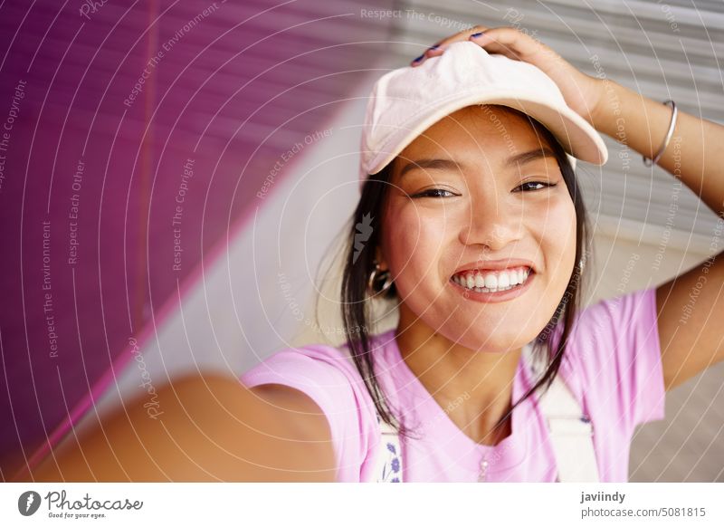 Cheerful Asian woman taking selfie on street smile style happy urban appearance touch head cap female young ethnic asian dark hair positive contemporary glad