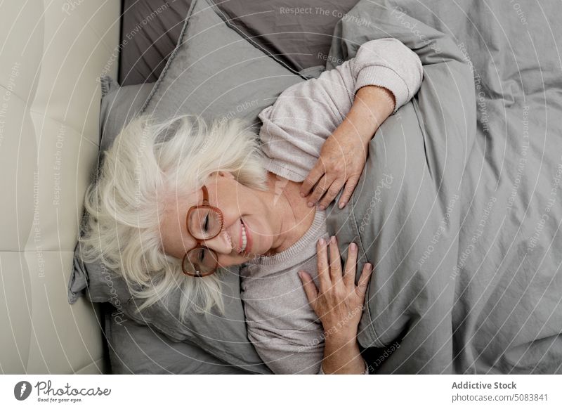 Elderly woman in eyeglasses lying on bed pensive thoughtful pensioner blanket bedclothes comfort rest bedroom relax home female senior sweater soft chill