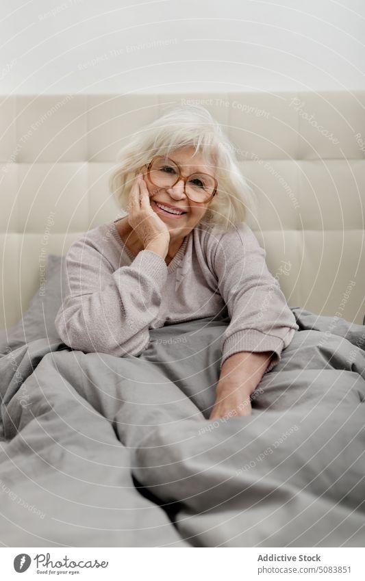 Elderly woman in eyeglasses sitting on bed happy pensioner blanket bedclothes comfort rest bedroom relax home female senior sweater soft chill domestic alone