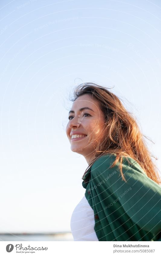 Beautiful young woman smiling with blue sky on the background. Caucasian woman with brown hair smiling and looking ahead wearing green outfit. Lifestyle candid female portrait.