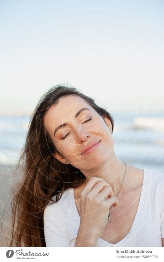 young adult caucasian woman smiling peacefully on the beach near sea with eyes closed. Female candid portrait of relaxed person. Caucasian woman with brown hair.