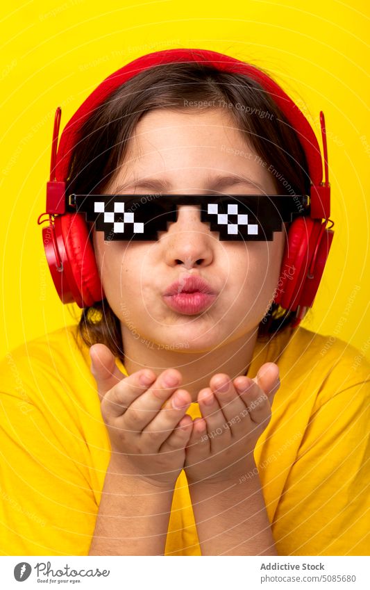 Cute girl sending air kiss while standing on yellow background kid gesture headphones music pixel glasses love cute adorable sweet schoolgirl child device