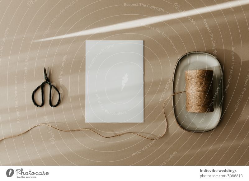 Stylish flat lay with a white blank paper greeting card, a black metal scissors, and a roll of twine string. Top view, copy space image. Creative message concept on beige background. Autumn colors.