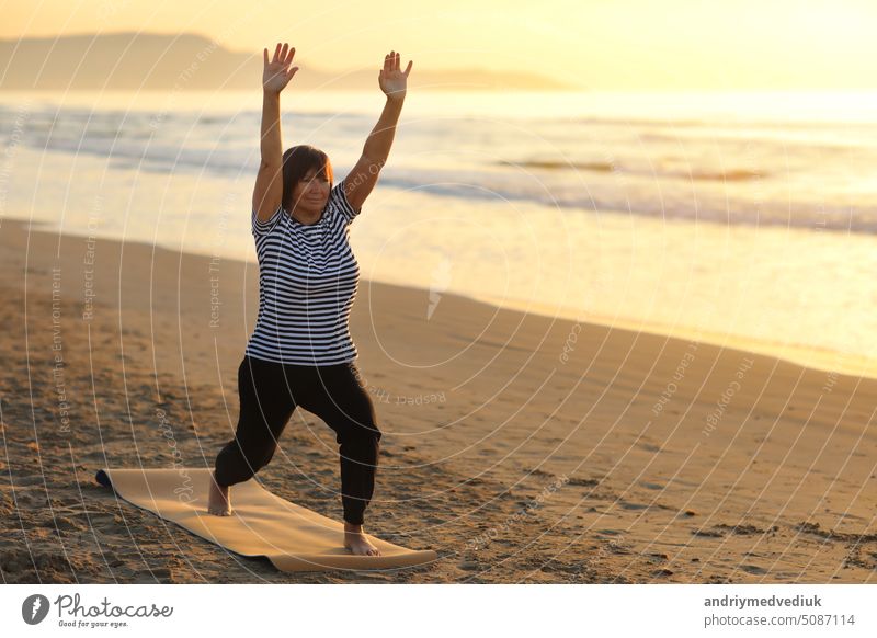 Mature female takes care of body and does yoga by the sea outside on sunrise. Female dressed the sport wear and do sport exercises in sandy beach. Meditation, yoga and relaxation concept.