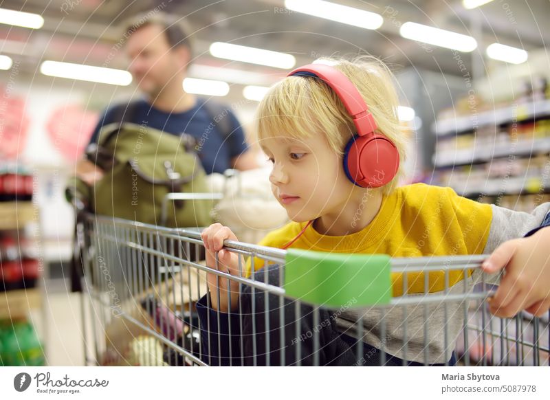 Cute preschooler boy with headphone and player is sitting in a shopping cart at a food store or supermarket. A child is listening to music or an audiobook while his parents buying groceries.